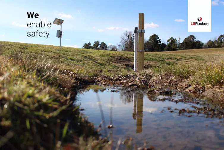 A flood monitoring pole is in the mid ground of the image, there is a small area of water or wetland in front of the pole. A slight grassy hill goes up to an unseen rail track but there is a solar panel atop the hill. There are trees in the distance. Text on the image says