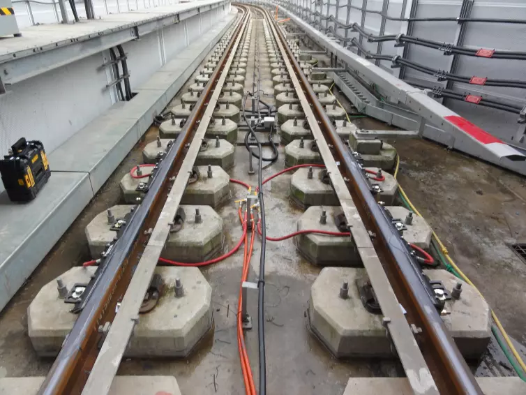 A railroad track with concrete slabs where the rail is supported by fasteners.  Bars with orange hoses are attached to the rails to product to the Top of the Rail.